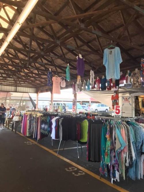 A bustling market with clothing racks and hanging garments under a wooden roof, shoppers browsing in the background.