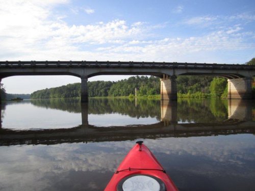 A red kayak on calm water, with a bridge reflected above and lush greenery lining the riverbanks under a blue sky.