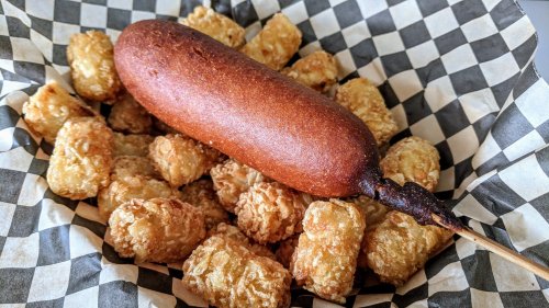 A crispy corn dog on a stick surrounded by golden tater tots in a checkered paper basket.