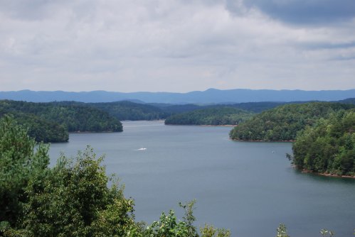 A serene lake surrounded by lush green hills and distant mountains under a cloudy sky.
