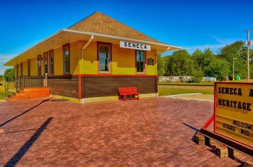 Historic yellow train station with a red bench, surrounded by a brick pathway and greenery, labeled "Seneca."