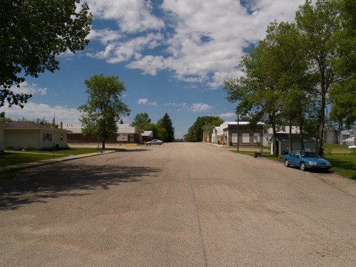 A quiet street lined with trees and buildings under a blue sky with scattered clouds. A blue car is parked on the side.