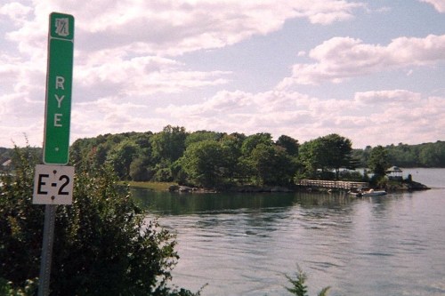 A green road sign for Rye near a calm waterway, with trees and a small dock in the background under a cloudy sky.