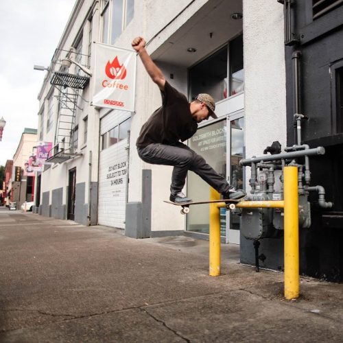 A skateboarder performs a trick on a yellow pole in an urban setting, with a coffee shop in the background.