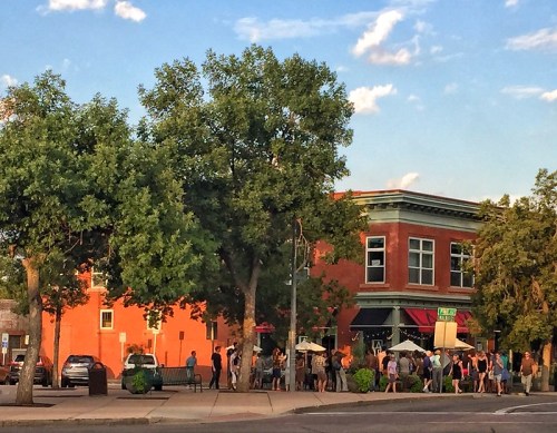 A busy street scene with a long line of people outside a red brick building under a blue sky with scattered clouds.