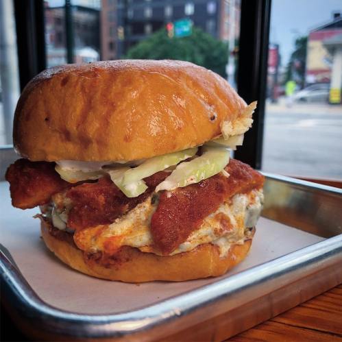 A crispy fried chicken sandwich with lettuce on a soft bun, served on a metal tray by a window.