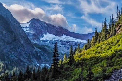 A scenic mountain landscape featuring a glacier, lush green trees, and a cloudy sky.