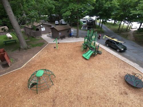 Aerial view of a playground with green equipment, surrounded by trees and a gravel path near parked cars and cabins.