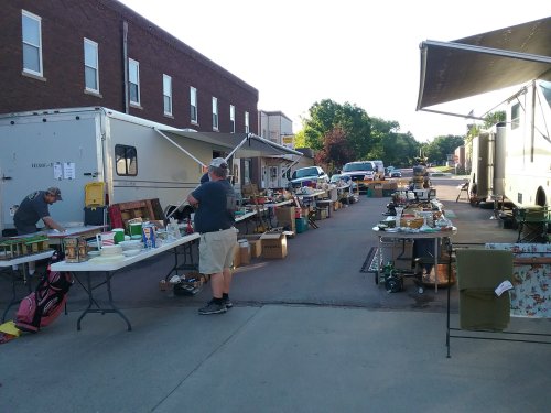 A bustling outdoor market with tables of various items for sale and vendors setting up in a sunny street.