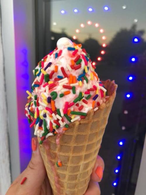 A hand holds a colorful ice cream cone topped with pink ice cream and vibrant sprinkles against a lit background.