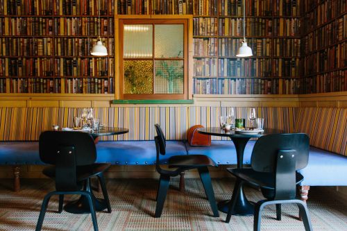Cozy restaurant interior with striped seating, wooden tables, and a bookshelf wallpaper. Soft lighting enhances the ambiance.