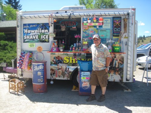 A man stands in front of a colorful shave ice truck, showcasing various flavors and treats. Bright signage and decorations are visible.