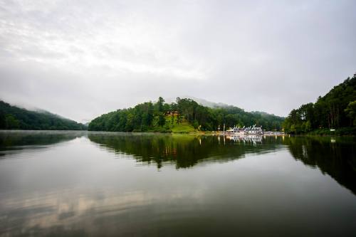 A serene lake surrounded by lush green hills, reflecting the cloudy sky and a distant building on the shore.