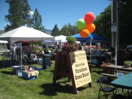 A vibrant outdoor market with tents, colorful balloons, and a sign for "Crazy Horse" offering furniture and home decor.
