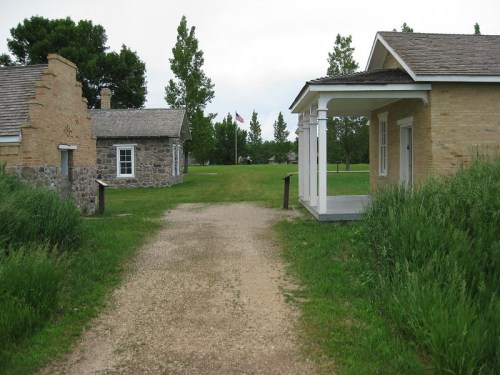 A gravel path leads between two historic buildings, surrounded by green grass and trees, with flags in the background.