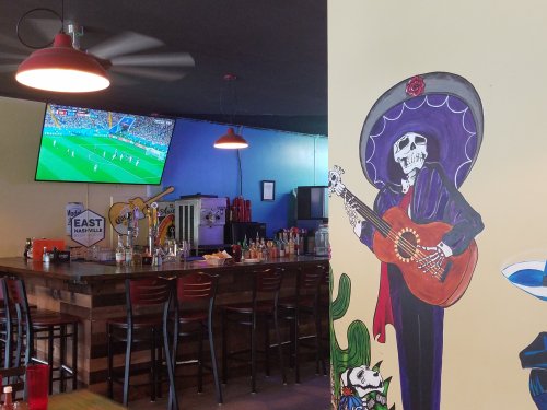 A vibrant bar scene with a mural of a skeleton playing guitar and a TV showing a soccer match in the background.