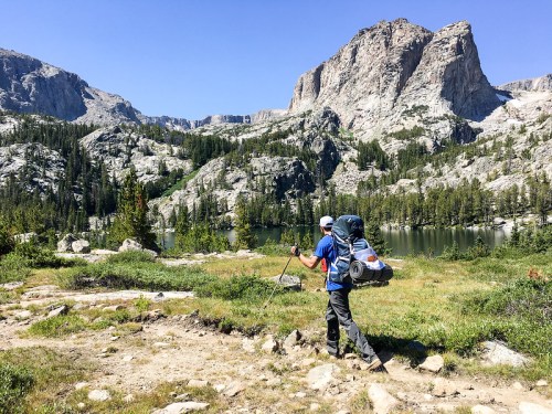 A hiker with a large backpack walks along a trail by a serene lake, surrounded by mountains and greenery.