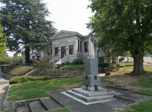 A historic library building with a statue in front, surrounded by greenery and trees on a sunny day.