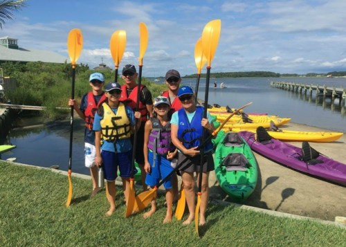 A group of six people in life jackets holding paddles, standing by a dock with colorful kayaks in the background.