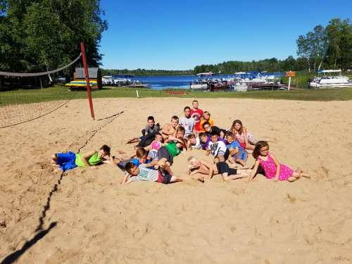 A group of children lying on a sandy beach near a lake, with boats and trees in the background.