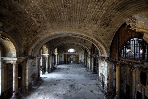 An expansive, abandoned interior with arched ceilings, crumbling walls, and scattered debris. Natural light filters through windows.
