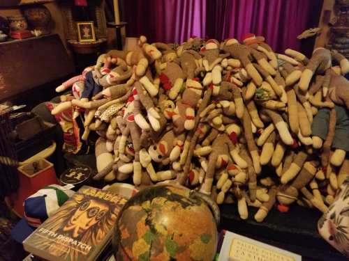 A large pile of vintage sock monkeys on a table, surrounded by books and a globe, with a colorful curtain in the background.
