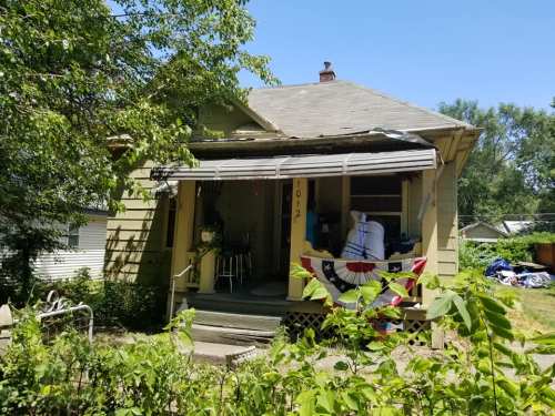 A green house with a porch, featuring a striped awning and patriotic decorations, surrounded by greenery.