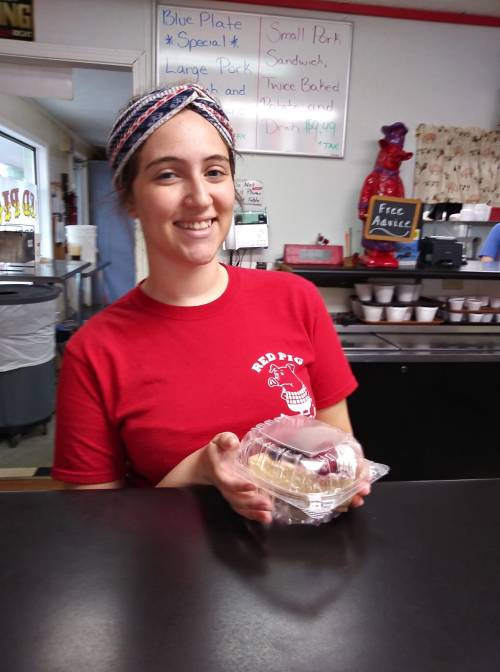 A smiling employee in a red shirt holds a dessert container at a diner, with a menu board in the background.