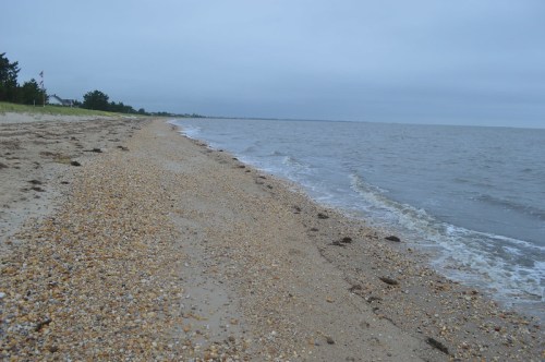 A sandy beach with pebbles and gentle waves under a cloudy sky, stretching along the shoreline.