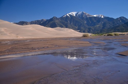 A serene landscape featuring sandy dunes, a river, and snow-capped mountains under a clear blue sky.