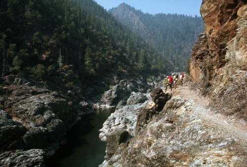 Hikers traverse a rocky path along a river, surrounded by dense forested mountains under a clear blue sky.