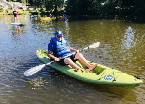 A person in a blue life jacket sits in a green kayak on a calm lake, with others paddling in the background.