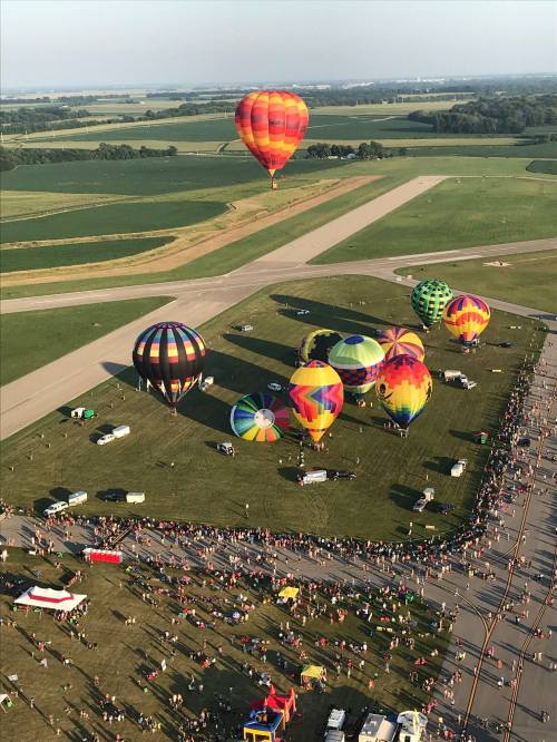 Aerial view of colorful hot air balloons over a grassy field, with a crowd gathered below and fields in the background.