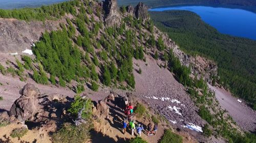 Aerial view of a rocky mountain landscape with hikers near a lake surrounded by dense green forest.