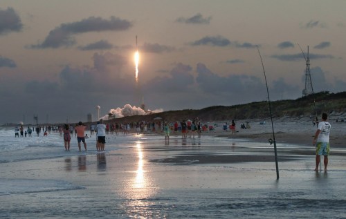 A rocket launches into the sky at sunset, with beachgoers and fishermen watching from the shore.