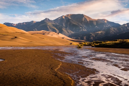 A serene landscape featuring sand dunes, a river, and mountains under a clear blue sky.