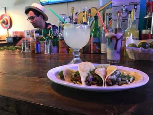 A plate of tacos sits on a wooden bar, with a margarita and a bartender in the background surrounded by drinks.