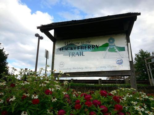 Sign welcoming visitors to the Huckleberry Trail, surrounded by colorful flowers and a cloudy sky.