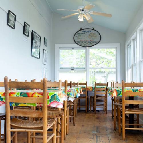 A bright dining area with wooden tables and chairs, featuring colorful tablecloths and large windows.