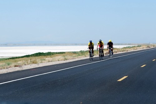 Three cyclists ride along a deserted road beside a vast, white salt flat under a clear blue sky.