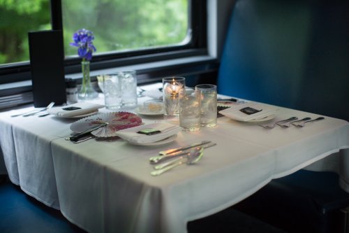 A elegantly set dining table with a white tablecloth, candles, glassware, and a small vase of flowers by a window.