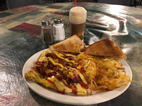 A plate with a loaded omelet topped with ketchup and mustard, hash browns, toast, and a frosty drink on a colorful table.