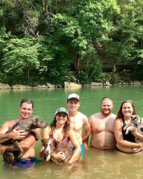 A group of five people wading in a river, each holding a dog, surrounded by lush greenery.