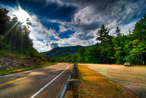 A scenic road curves through lush greenery under a bright sky, with a parking area and distant mountains visible.