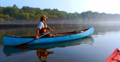 A person paddles a blue kayak on a calm river surrounded by misty trees in the early morning light.