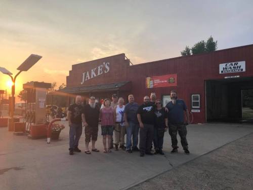 A group of people stands outside a red building labeled "Jake's," with a car wash sign and sunset in the background.
