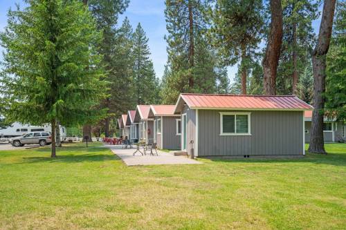 Row of small gray cabins with red roofs, surrounded by tall trees and green grass on a sunny day.