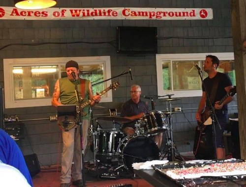 A band performs at a campground, featuring a guitarist, drummer, and bassist in a rustic indoor setting.
