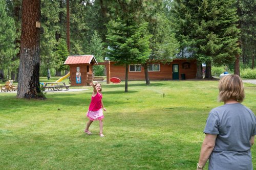 A girl in a pink dress throws a frisbee in a grassy area, while an adult watches nearby, surrounded by trees and cabins.