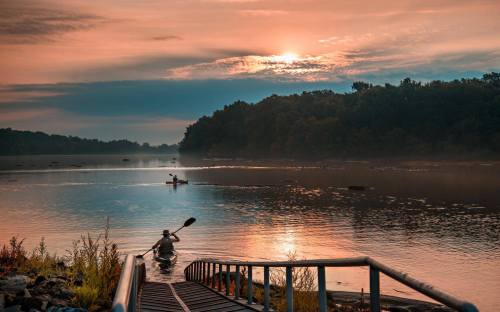 A serene river scene at sunrise, with a person kayaking and another in the distance, surrounded by misty trees.